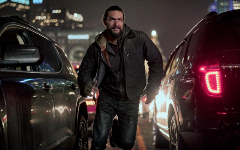 Jason Momoa corre pela rua entre carros em cena do filme Justiça em Família