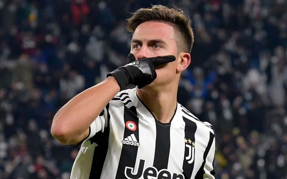 Jogador Dybala, da Juventus, veste uniforme branco com listras pretas e comemora gol com a mão no rosto