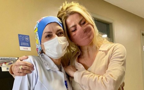 Karina Bacchi de máscara marrom, camisa bege, de olhos fechados, abraçada á enfermeira, de touca azul
