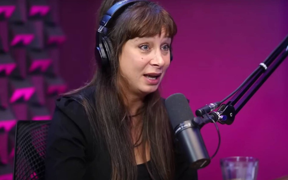 A atriz Katiuscia Canoro com fones de ouvido, de boca aberta, em frente a microfone em estúdio de podcast