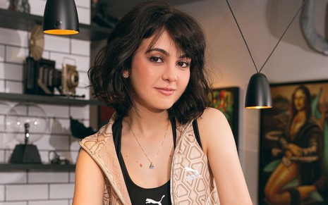 Klara Castanho usa blusa preta e colete bege: ela posa em um fundo com prateleira com quadros e luminárias atrás