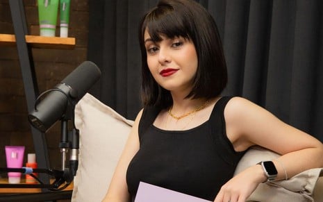 Klara Castanho usa roupa preta e batom vermelho e posa em um estúdio com microfone à sua frente