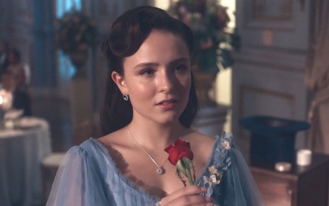 Larissa Manoela em cena de Além da Ilusão: atriz está caracterizada com vestido azul, cabelo preso e segura rosa vermelha