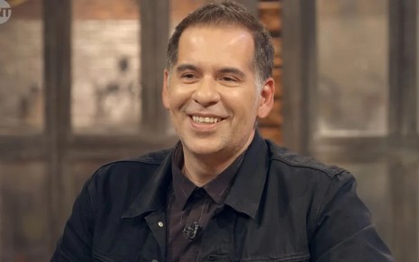 Leandro Hassum com um terno e camisa preta, sorrindo para a câmera, durante o programa Tá Pago, da TNT