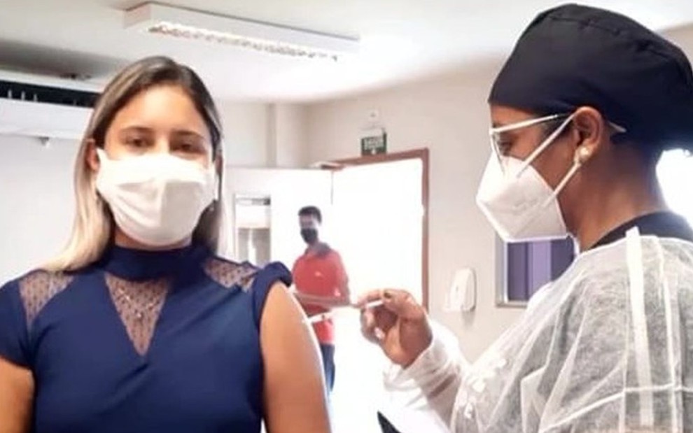 Lena Mendonça, loira, de blusa azul, tomando a vacina no braço esquerdo