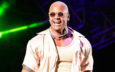 Léo Santana está sorrindo em cima do palco; ele usa óculos escuros, camiseta branca e ua blusa de botão por cima