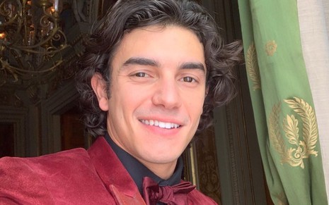 Leonardo Zanchin dá sorriso de lado, vestindo paletó e gravata vermelha, em registro nos bastidores de Quanto Mais Vida, Melhor!