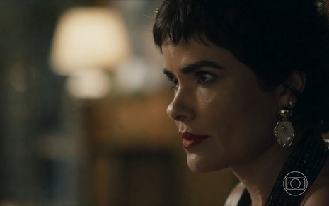 Em cena de Travessia, Vanessa Giácomo está de lado, olhando para alguém a sua frente, usando um brinco com pedra de cor perolada e batom vermelho