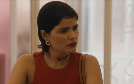 Em cena de Travessia, Vanessa Giácomo está com a expressão de surpresa conversando com alguém; ela usa camiseta vermelha