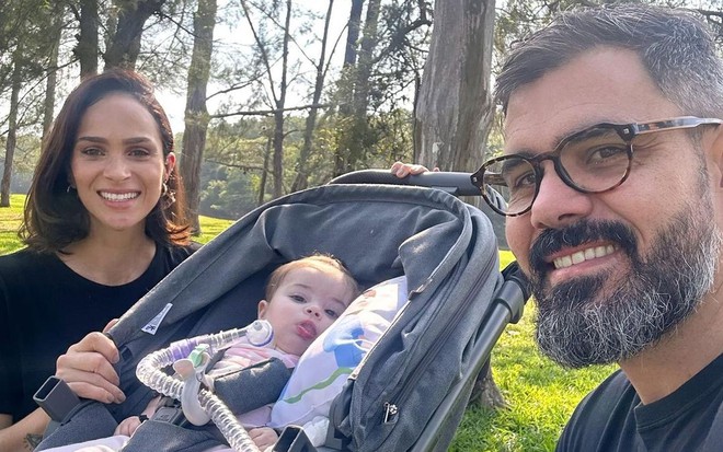 Letícia e Juliano Cazarré sorriem em um parque, com Maria Guilhermina entre eles em um bercinho