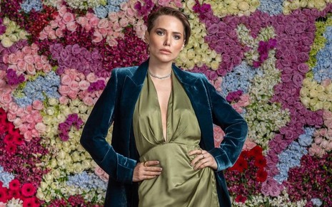De vestido verde e blazer azul, Leticia Colin posa com um painel de flores ao fundo