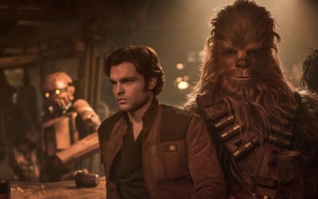 O ator Alden Ehrenreich como o personagem Han Solo e Joonas Suotamono como Chewbacca no filme Han Solo: Uma História Star Wars; os personagens aparecem no meio de um bar com um robô ao fundo e à direita