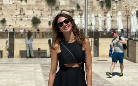 Luciana Gimenez usa um vestido preto com um recorte logo abaixo dos seios; ao fundo, é possível ver o Muro das Lamentações