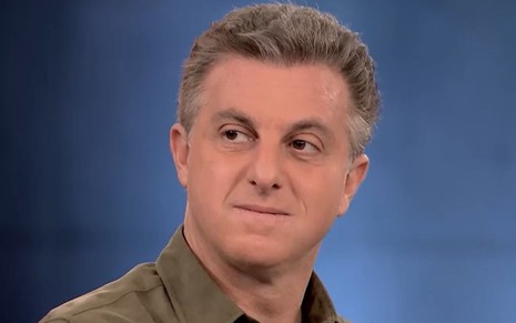 Luciano Huck usa camisa verde e olha para o lado enquanto ouve pessoa falar durante programa na Globo