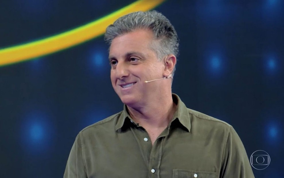 Luciano Huck de perfil durante gravação do Caldeirão. Apresentador veste camisa verde e sorri.