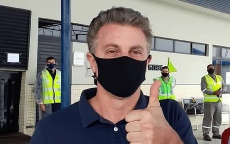 Luciano Huck com uma camisa azul e usando uma máscara preta, atendendo fãs no aeroporto de Campina Grande, na Paraíba. Ele faz o sinal de positivo.