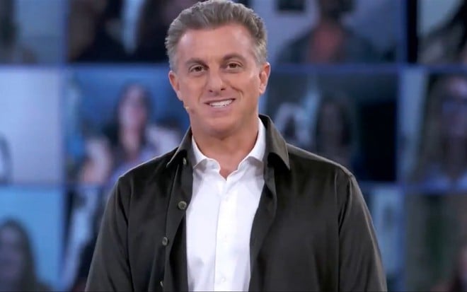 Luciano Huck, vestido com uma camiseta marrom, no estúdio de fundo azul de seu programa na Globo que vai ao ar nas tardes de sábado