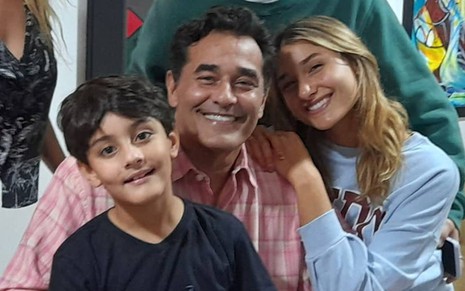 Luciano está sorrindo, veste camiseta rosa e está ao lado de um de seus filhos, que veste camiseta preta e está sorrindo; ao lado de Luciano está Sasha, que sorri e veste camiseta azul