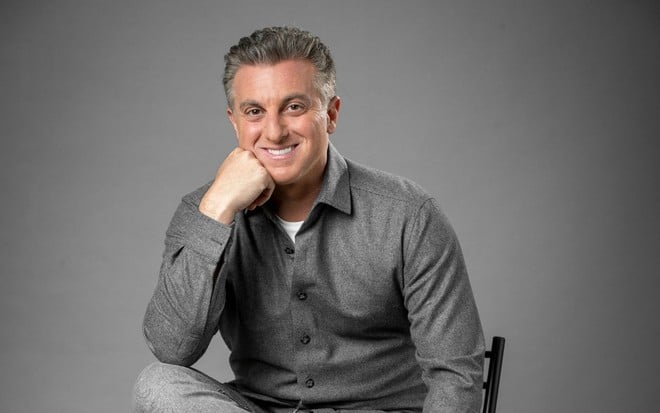 Luciano Huck usa uma camisa cinza e posa para foto com a mão direita apoiando o rosto