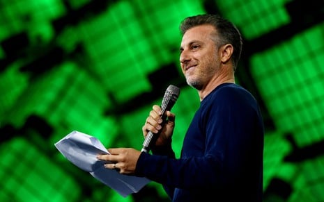 O apresentador Luciano Huck na abertura da Web Summit Rio; ele usa camiseta escura; ao fundo, painel verde