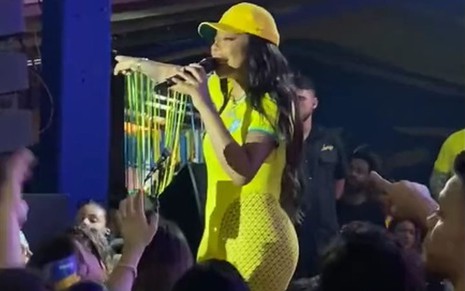 De boné e macacão verdade e amarelo, Ludmilla está em cima do palco, com o dedo em riste, falando ao microfone
