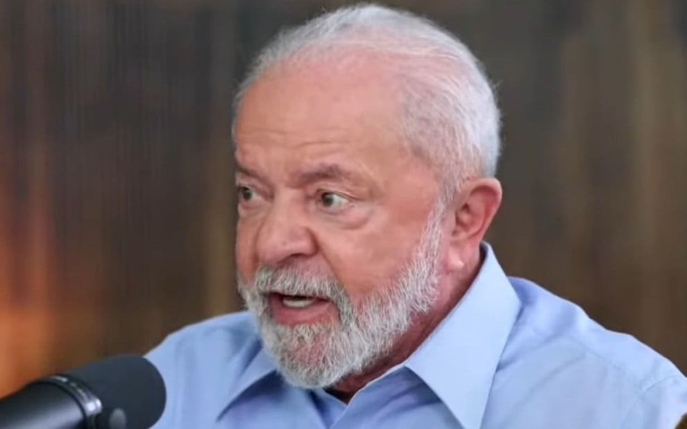Luiz Inácio Lula da Silva usa camisa azul e está com expressão séria falando num microfone estilo podcast