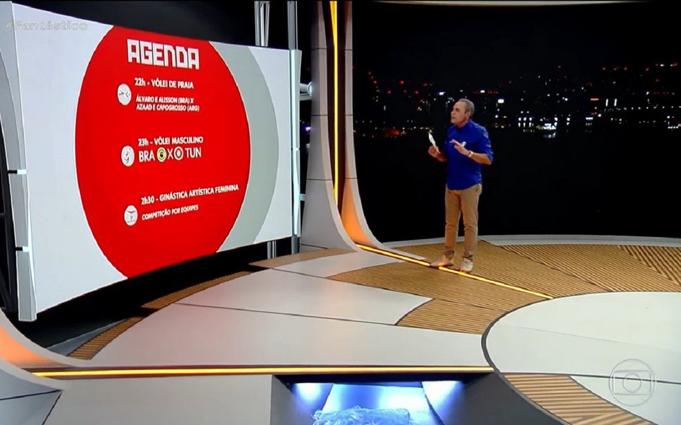 Luis Roberto nos estúdios olímpicos da Globo no Rio de Janeiro. O estúdio tem iso marrom e branco, com um telão grande e fundo escuro.