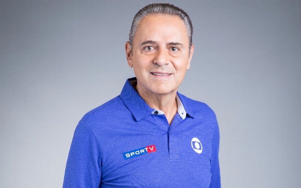 Luis Roberto com uma camisa azul, num fundo cinza, sorrindo para a câmera e usando o uniforme do Esporte da Globo