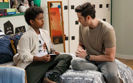 Nando (Lukas Cabral) conversa com Fred (Velson D'Souza); ambos estão sentados em cama