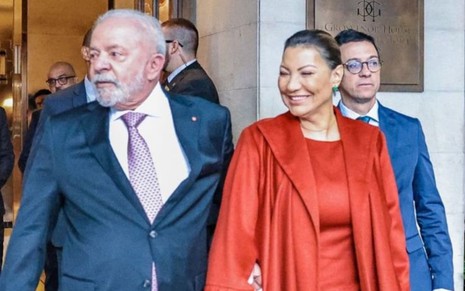 Lula segura a cintura de Janja enquanto os dois andam pelas ruas; ele usa um terno preto e uma gravata rosada; ela, vestido e casaco laranjas