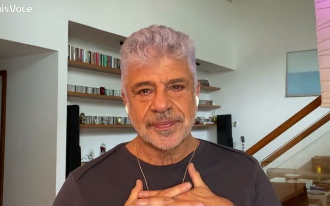 Lulu Santos em sua casa vestindo uma camiseta de manga longa preta e um colar dourado. O cantor está com lágrimas nos olhos, uma feição emocionada e as duas mãos no peito. No fundo é possível observar paredes brancas, uma estante e uma escada.