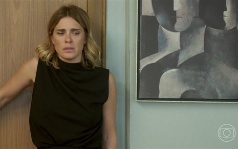 Em cena de Vai na Fé, Carolina Dieckmann está vestindo roupa preta e está encostada em uma parede com a expressão de tristeza