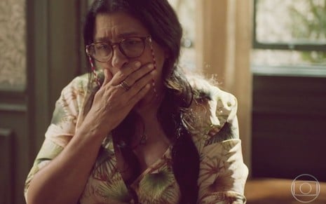 A atriz Regina Casé como Lurdes em Amor de Mãe; ela está olhando para baixo com a mão na boca e cara de tristeza