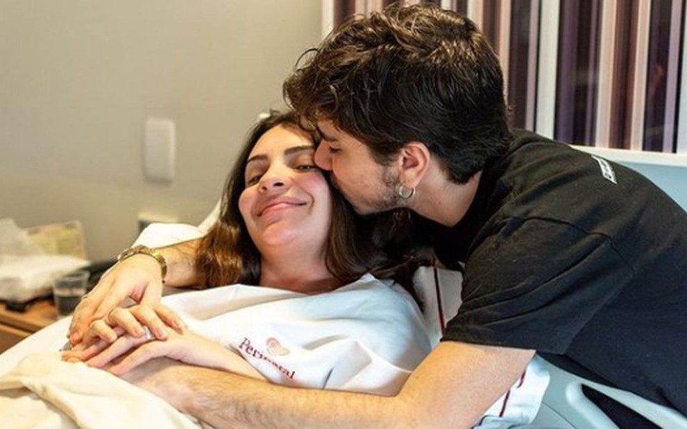 Mabel Calzolari com sorriso contido, deitada na cama de hospital, ganhando beijo de João Fernandes