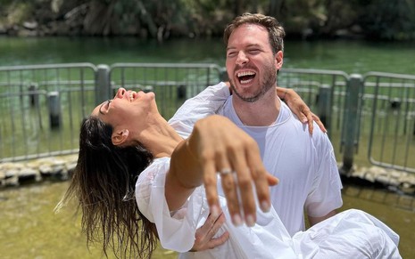Maíra Cardi e Thiago Nigro estão vestidos de branco, molhados no rio. Ela exibe aliança de noivado e está no colo do noivo