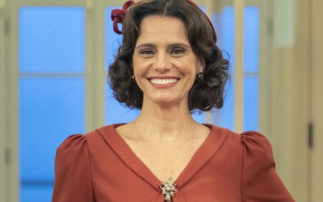 Na imagem, a atriz Malu Galli está caracterizada como Violeta, sua personagem na novela Além da Ilusão. Com o cabelo curto e batom claro, ela está vestida com uma roupa vermelha e tem um acessório no cabelo.