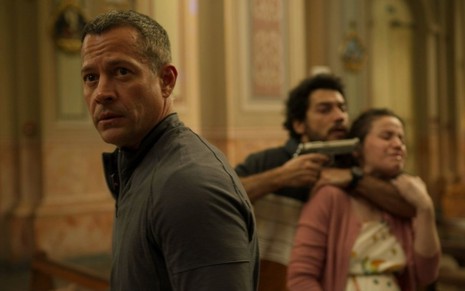 Malvino Salvador à frente e ator ao fundo segura um revólver na cabeça de uma mulher, também atriz