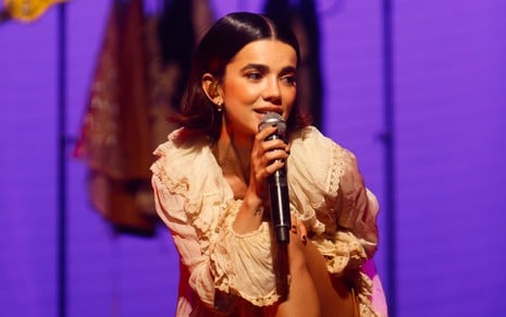 A cantora Manu Gavassi está em um palco com uma roupa clara e segura microfone pertinho da boca