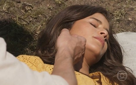 A atriz Isadora Cruz como Candoca em Mar do Sertão; ela está deitada sobre a grama e dormindo enquanto a mão de José (Sergio Guizé) se aproxima de seu rosto