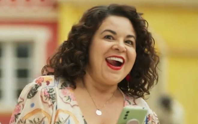 A atriz Suzy Lopes como Cira em Mar do Sertão; ela está olhando para a frente enquanto segura um celular com a boca aberta e cara de feliz após conseguir o que queria
