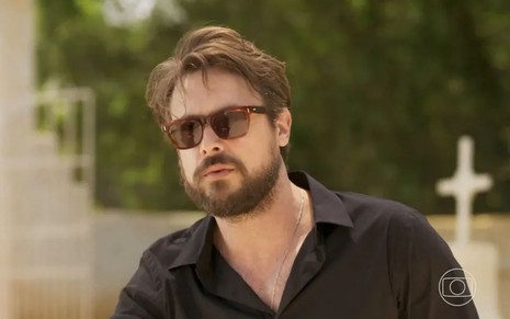 O ator Sergio Guizé como José em Mar do Sertão; ele está agachado, usando um óculos de sol, olhando para frente com cara séria