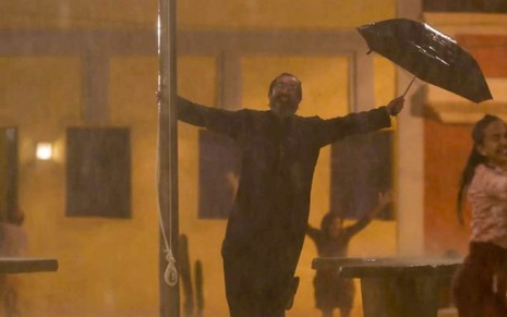O ator Nanego Lira como Padre Zezo em Mar do Sertão; ele está escorado em um poste segurando um guarda-chuva na outra mão e sorrindo em meio à chuva