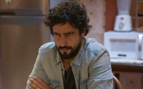 O ator Renato Góes como Tertulinho em Mar do Sertão; ele está sentado a uma mesa, com os braços cruzados e as mãos sobre os antebraços opostos, enquanto olha para frente com cara de maquiavélico