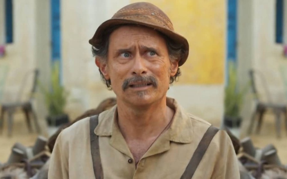 Enrique Diaz caracterizado como Timbó, ele veste roupas beges e surradas e tem a expressão séria em cena de Mar do Sertão