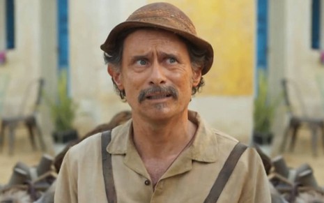 Enrique Diaz caracterizado como Timbó, ele veste roupas beges e surradas e tem a expressão séria em cena de Mar do Sertão
