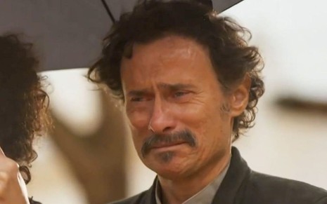 O ator Enrique Diaz como Timbó em Mar do Sertão; ele está debaixo de um guarda-chuva, olhando para o lado com cara de choro