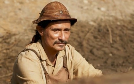 O ator Enrique Diaz como Timbó em Mar do Sertão; ele está sentado, olhando para a frente com cara de pensativo