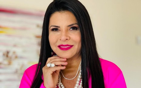 A apresentadora Mara Maravilha com blusa rosa, colares e anéis de pérolas, sorrindo para foto