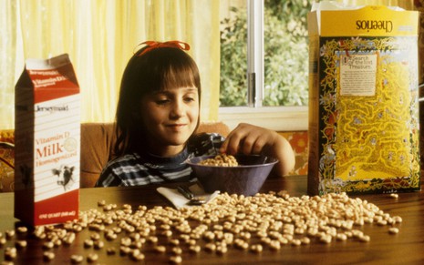 Mara Wilson sentada na frente de um monte de cereal espalhado em cena de Matilda
