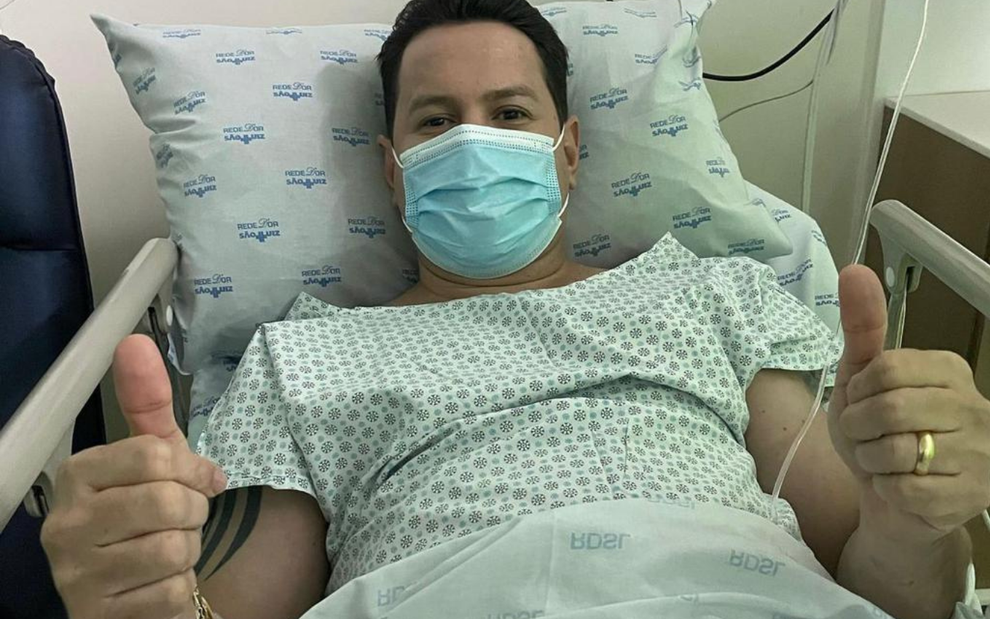 Marcão do Povo está em uma cama de hospital; ele usa máscara e faz sinal de joinha